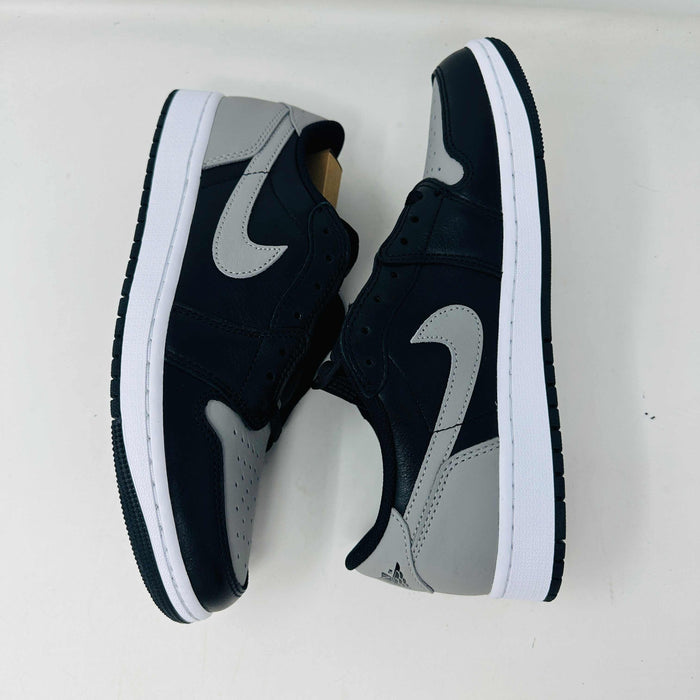 A pair of Jordan 1 Low OG Shadow (2024) sneakers, in black and gray, rests atop a gray shoe box with a large black Nike logo. Beside the box is a small brown cardboard package featuring a cutout revealing shoelaces inside.