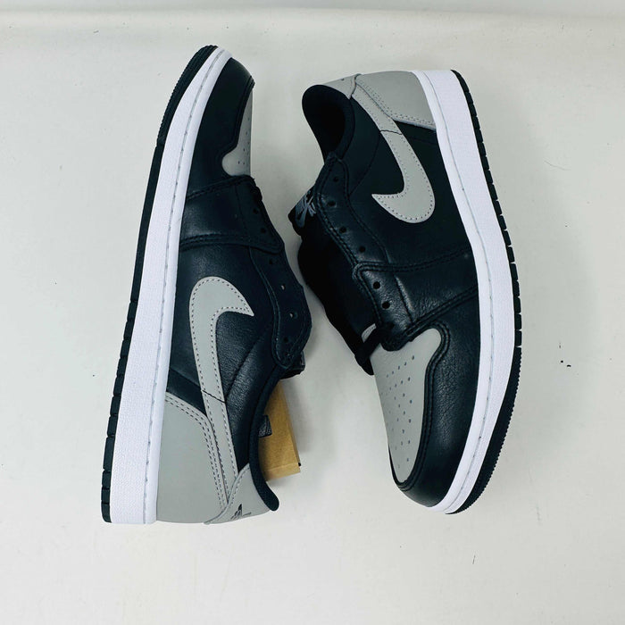 A pair of Jordan 1 Low OG Shadow (2024) sneakers, in black and gray, rests atop a gray shoe box with a large black Nike logo. Beside the box is a small brown cardboard package featuring a cutout revealing shoelaces inside.