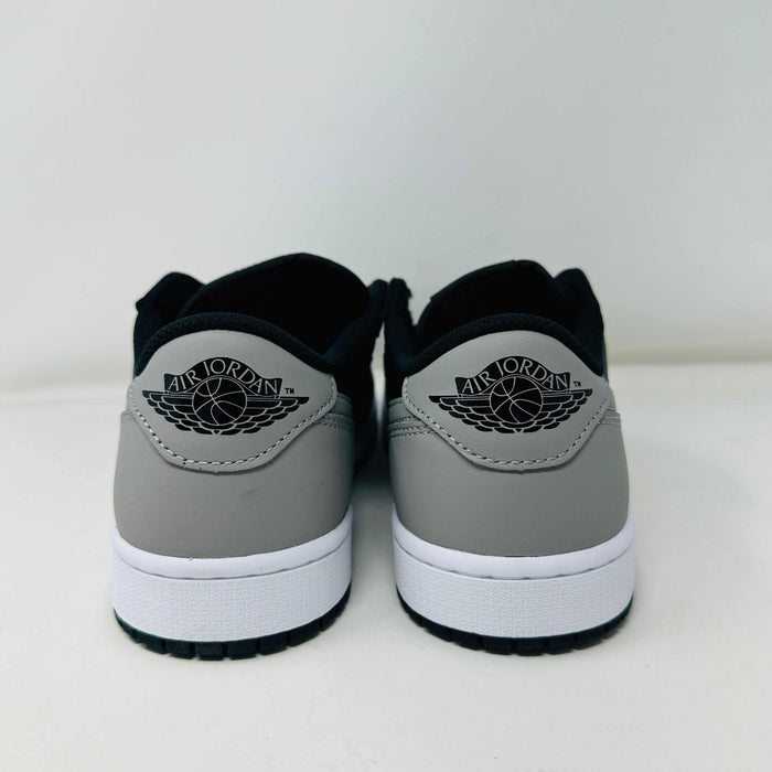 A pair of Jordan 1 Low OG Shadow (2024) sneakers, in black and gray, rests atop a gray shoe box with a large black Nike logo. Beside the box is a small brown cardboard package featuring a cutout revealing shoelaces inside.
