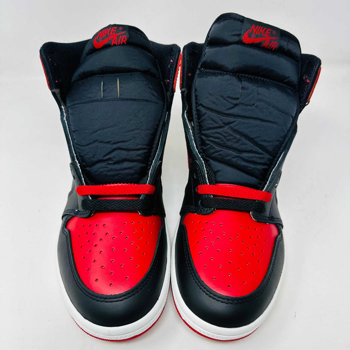 A Jordan 1 Retro High 85 OG Bred (2025) sneaker by Jordan sits on its matching shoebox, showcasing the iconic swoosh and Air Jordan logo. Its styled in black and red with a white midsole, bold red laces, and detailed black accents.