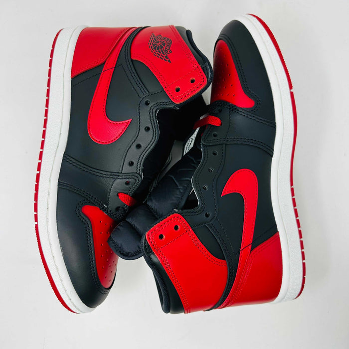 A Jordan 1 Retro High 85 OG Bred (2025) sneaker by Jordan sits on its matching shoebox, showcasing the iconic swoosh and Air Jordan logo. Its styled in black and red with a white midsole, bold red laces, and detailed black accents.
