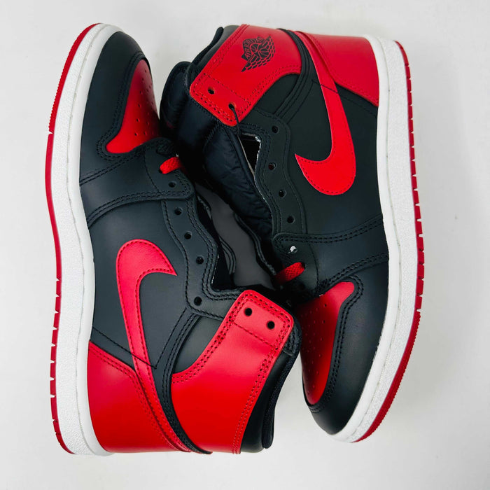A Jordan 1 Retro High 85 OG Bred (2025) sneaker by Jordan sits on its matching shoebox, showcasing the iconic swoosh and Air Jordan logo. Its styled in black and red with a white midsole, bold red laces, and detailed black accents.
