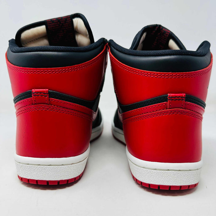 A Jordan 1 Retro High 85 OG Bred (2025) sneaker by Jordan sits on its matching shoebox, showcasing the iconic swoosh and Air Jordan logo. Its styled in black and red with a white midsole, bold red laces, and detailed black accents.