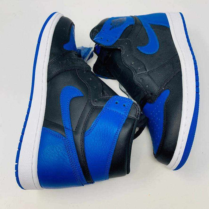 A pair of brand new black and blue Jordan 1 Royal 2017 high-tops are showcased on a black and red Nike box, with a small plastic bag holding authentic blue laces attached to one shoe.