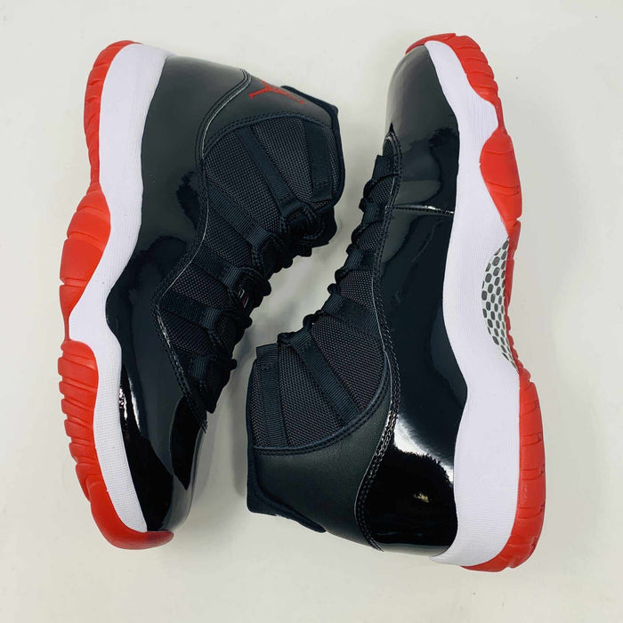 The brand new Jordan 11 Bred 2019 features a black and red high-top design with glossy accents, striking red soles, and a bold red Jumpman logo. It sits atop an orange and black Nike shoebox, exuding style and performance.