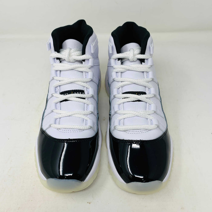 A pair of authentic Jordan 11 Retro DMP Gratitude (2023) (GS) sneakers, featuring a glossy white and black finish with a gold logo, is placed on top of a gray Nike Air shoebox.