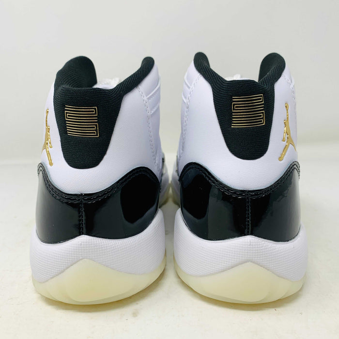 A pair of authentic Jordan 11 Retro DMP Gratitude (2023) (GS) sneakers, featuring a glossy white and black finish with a gold logo, is placed on top of a gray Nike Air shoebox.