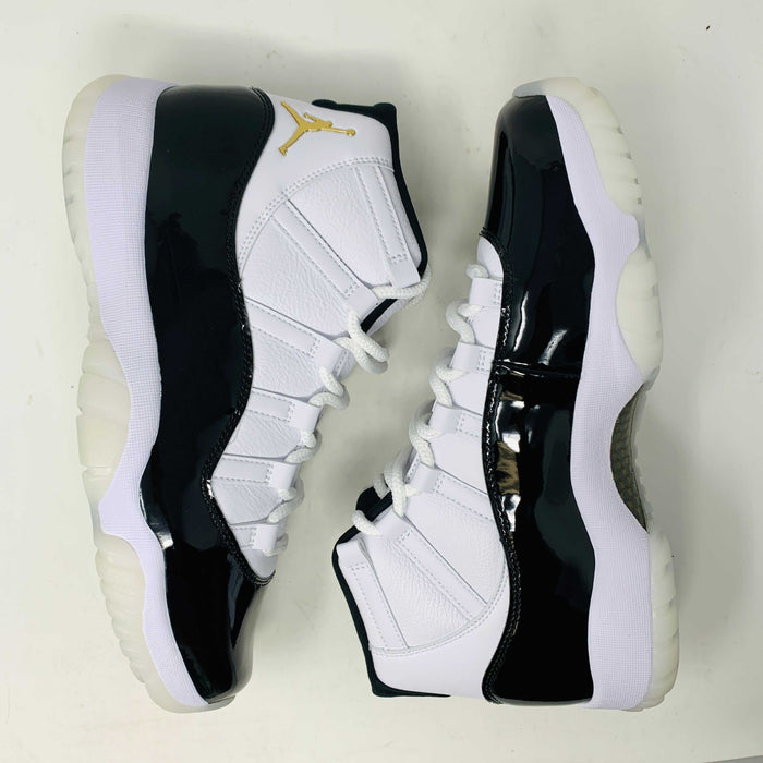 The Jordan 11 Retro DMP Gratitude (2023) features high-top sneakers in a white-black colorway with shiny accents. They have white laces, a crisp white midsole, and a translucent outsole, adorned with a small golden ankle logo and come in a sleek black box.