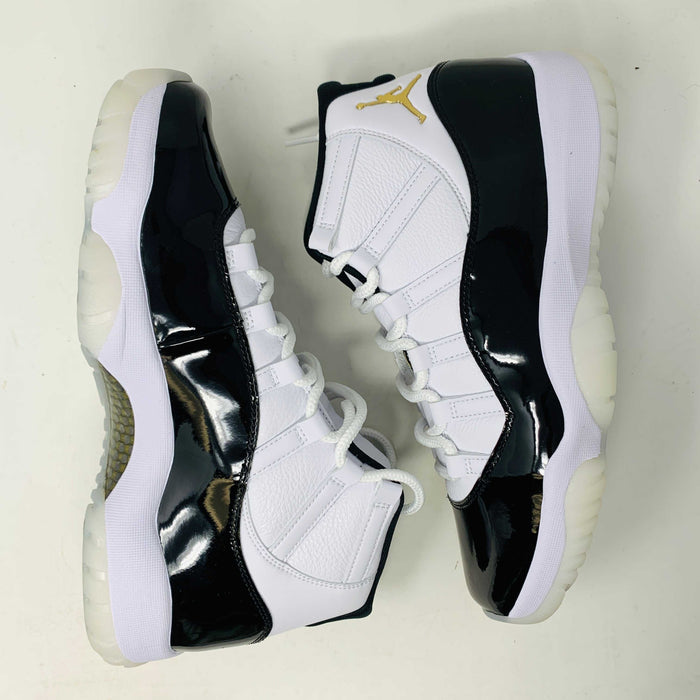 The Jordan 11 Retro DMP Gratitude (2023) features high-top sneakers in a white-black colorway with shiny accents. They have white laces, a crisp white midsole, and a translucent outsole, adorned with a small golden ankle logo and come in a sleek black box.