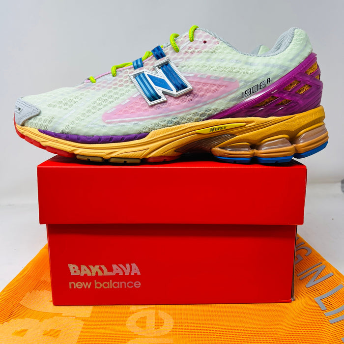 The New Balance 1906R Action Bronson Rosewater sneaker, boasting a light mesh upper and colorful sole with a bold blue N logo, rests on a pristine red Baylaya box. An orange surface partially peeks out under the box.
