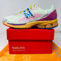 The New Balance 1906R Action Bronson Rosewater sneaker, boasting a light mesh upper and colorful sole with a bold blue N logo, rests on a pristine red Baylaya box. An orange surface partially peeks out under the box.