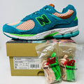 A pair of New Balance 2002R by Salehe Bembury, in colorful teal, pink, and green accents, sits atop a shoebox. Two extra lace sets—pink and green—are in front. The shoebox label details the size and style; the box is in good condition with a clean design.