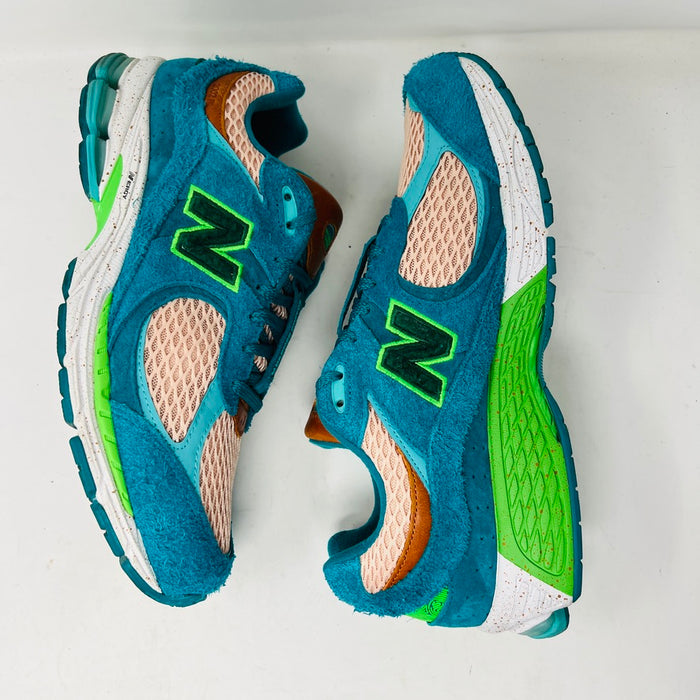 A pair of New Balance 2002R by Salehe Bembury, in colorful teal, pink, and green accents, sits atop a shoebox. Two extra lace sets—pink and green—are in front. The shoebox label details the size and style; the box is in good condition with a clean design.