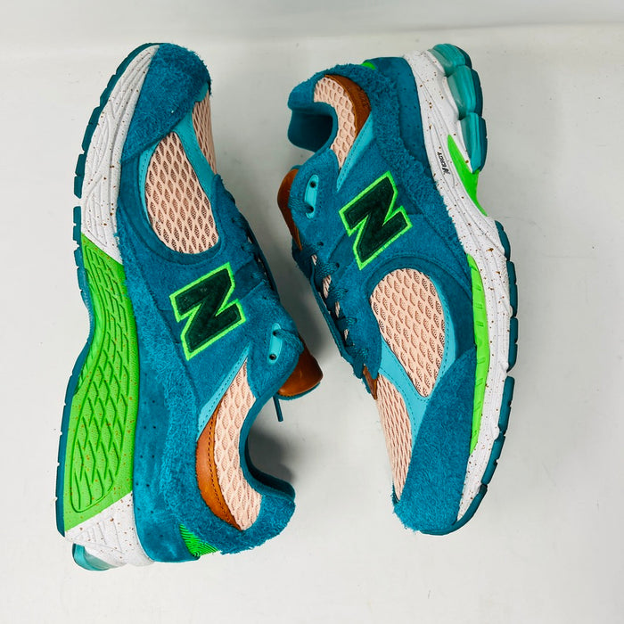A pair of New Balance 2002R by Salehe Bembury, in colorful teal, pink, and green accents, sits atop a shoebox. Two extra lace sets—pink and green—are in front. The shoebox label details the size and style; the box is in good condition with a clean design.