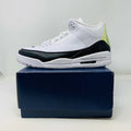 The Jordan 3 Fragment by Jordan, a white sneaker with black midsole and lime green heel detailing, is displayed on a closed blue shoebox.