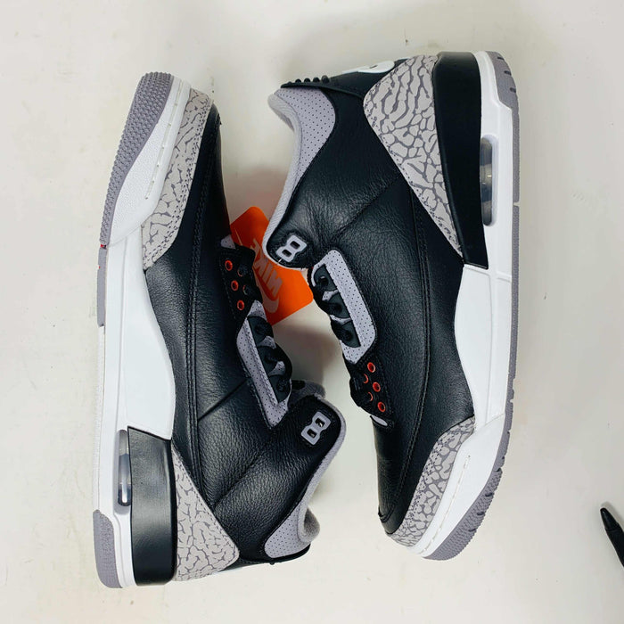 The Jordan 3 Retro OG Black Cement (2024) sneaker, featuring black and gray tones with red accents, is displayed on a sleek black shoebox. With textured details and visible air cushioning, an orange Nike Air tag authenticates this stylish addition to your collection.