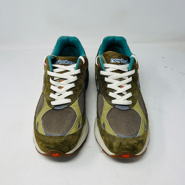 A New Balance 990v3 MiUSA Bodega Here To Stay sneaker in green and gray with white laces, detailed stitching, and side branding sits on a brown box featuring New Balance, Bodega, and 15 Years logos. This 2021 edition is a stylish wardrobe accessory.