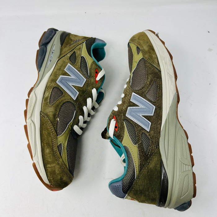 A New Balance 990v3 MiUSA Bodega Here To Stay sneaker in green and gray with white laces, detailed stitching, and side branding sits on a brown box featuring New Balance, Bodega, and 15 Years logos. This 2021 edition is a stylish wardrobe accessory.