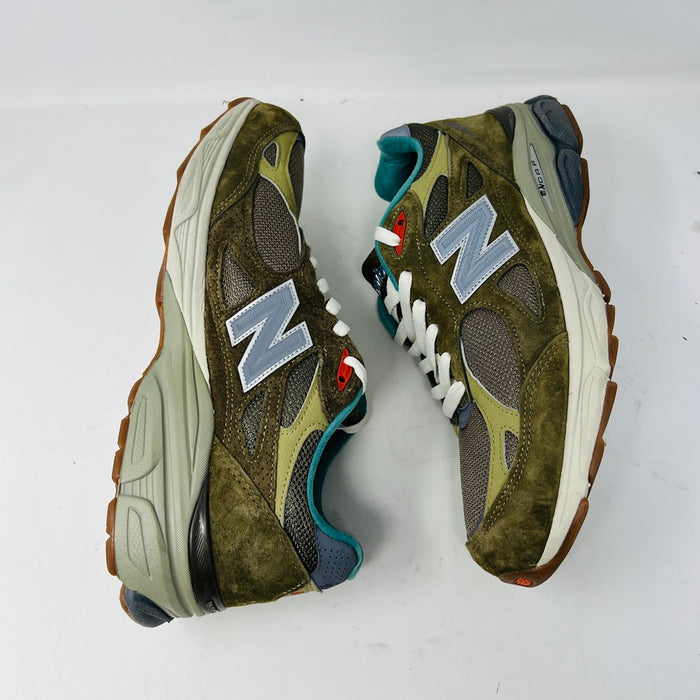 A New Balance 990v3 MiUSA Bodega Here To Stay sneaker in green and gray with white laces, detailed stitching, and side branding sits on a brown box featuring New Balance, Bodega, and 15 Years logos. This 2021 edition is a stylish wardrobe accessory.