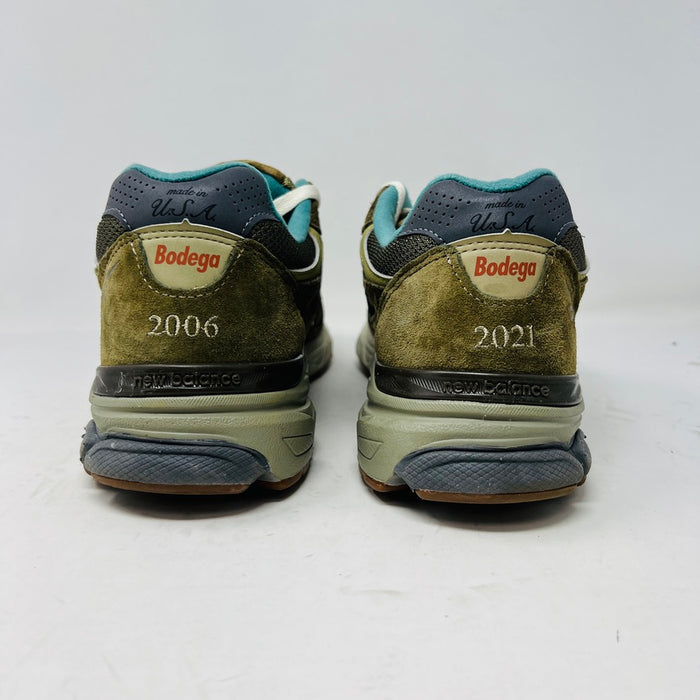 A New Balance 990v3 MiUSA Bodega Here To Stay sneaker in green and gray with white laces, detailed stitching, and side branding sits on a brown box featuring New Balance, Bodega, and 15 Years logos. This 2021 edition is a stylish wardrobe accessory.
