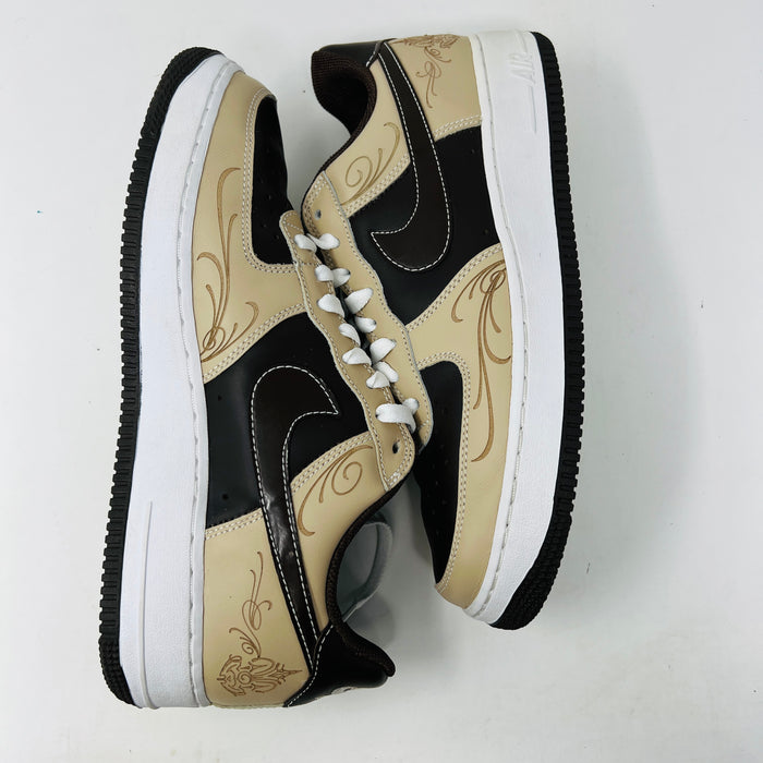 The Nike Air Force 1 Low Brown Pride sneakers feature beige and dark brown leather panels, white laces, and a white sole. They are shown from a top-down perspective in super clean condition on an immaculate white background.