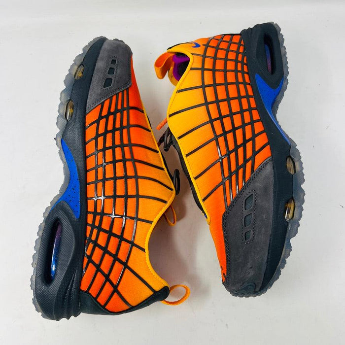 The Nike Air Max Sunder SP Kids of Immigrants Sunrise sneakers in bright orange and black feature a grid pattern with blue accents. An authentic swoosh and a circular K-I design mark the shoes and the orange box, highlighting their uniqueness.