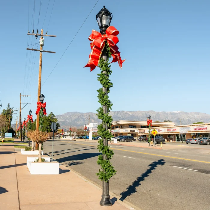 A black decorative street lamp on a city sidewalk wrapped in green garland with a large red structural bow with gold trim, photographed outdoors on a sunny day with mountains and a street in the background.