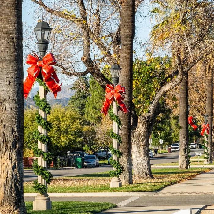 Outdoor street scene with tall lamp posts wrapped in green garland and large red structural bows, set among trees on a sidewalk near a road with parked cars.