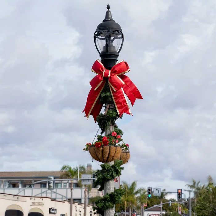 A black vintage-style street lamp decorated for the holidays with a large red structural bow, green garland, and two hanging flower baskets, photographed outdoors against a cloudy sky.