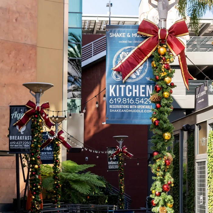 Outdoor storefront scene with multiple banners hanging from poles, decorated with large red structural bows, green garlands, pinecones, and red and gold ornaments. Buildings, windows, plants, and outdoor lighting are visible in the background.