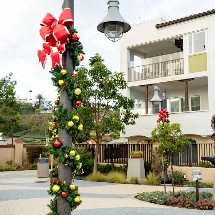 Christmas greenery garland wrapped around a street lamp post in an outdoor residential courtyard, decorated with a large red structural bow, red and gold ornaments, pinecones, and evergreen branches, with a modern apartment building in the background.