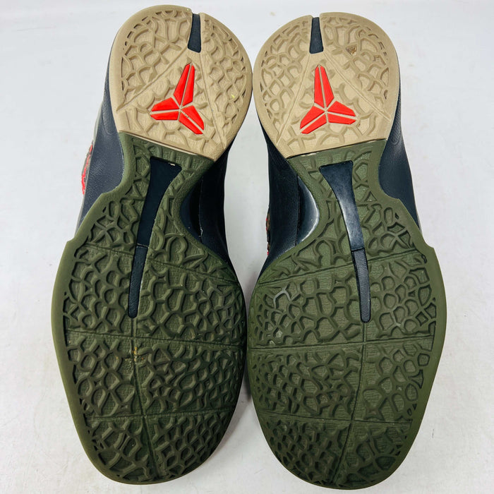 Close-up of a Nike Kobe 6 Protro Italian Camo (2024) (GS) shoe in black and dark olive with a red swoosh, resting on an orange Nike box. The 100% authentic shoe features a textured upper shown from the side, highlighting its design and striking color contrast.