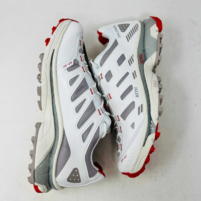 The Salomon XT-4K Kith Kithmas White Red sneakers rest on a black Kith box, with an extra laces bag. The shoes are 100% authentic, showcasing red accents and branding details.