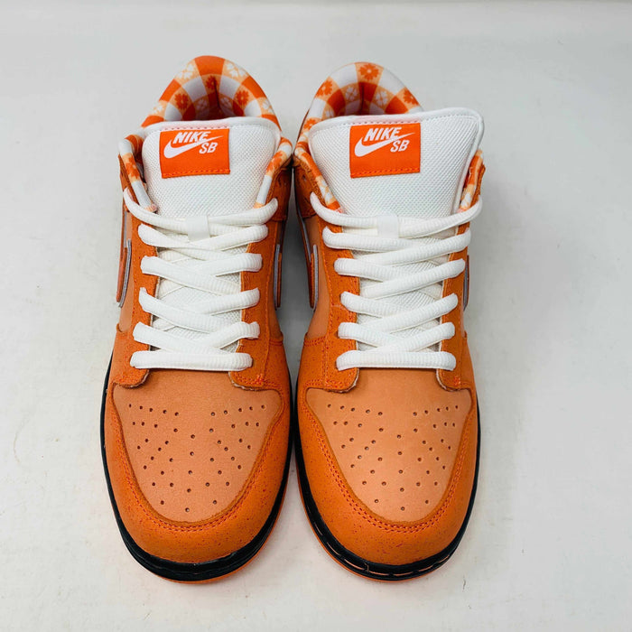 The authentic Nike SB Dunk Low Orange Lobster Concepts sneaker sits on a green and yellow Nike SB box. It features a white sole with black trim, an orange swoosh, and extra laces neatly positioned in front of the box.
