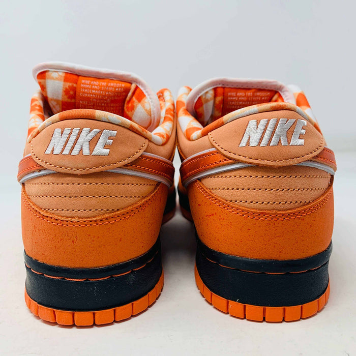 The authentic Nike SB Dunk Low Orange Lobster Concepts sneaker sits on a green and yellow Nike SB box. It features a white sole with black trim, an orange swoosh, and extra laces neatly positioned in front of the box.