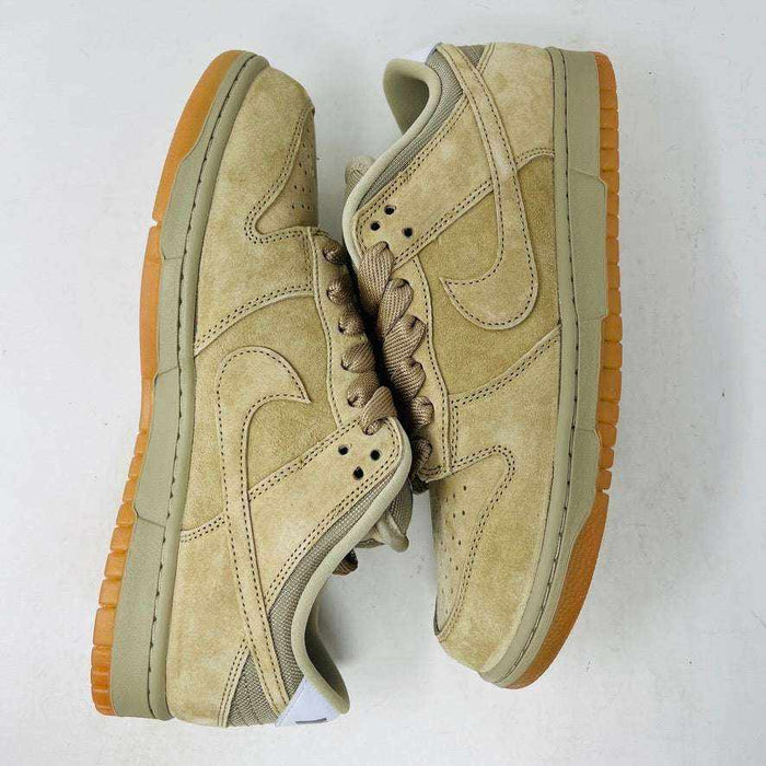 The Nike SB Dunk Low Pro B Parachute Beige, with a beige swoosh and gum sole, is displayed on a purple Nike SB box. New tan suede sneaker includes extra laces neatly rolled up in front against a plain white background.