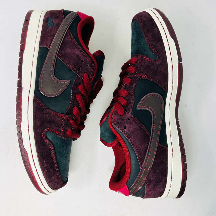 A BRAND NEW Nike SB Dunk Low Riot Skateshop sneaker featuring a black design with maroon accents, including a swoosh, laces, and a black sole with a white midsole, is displayed on top of its original white Nike SB shoebox.