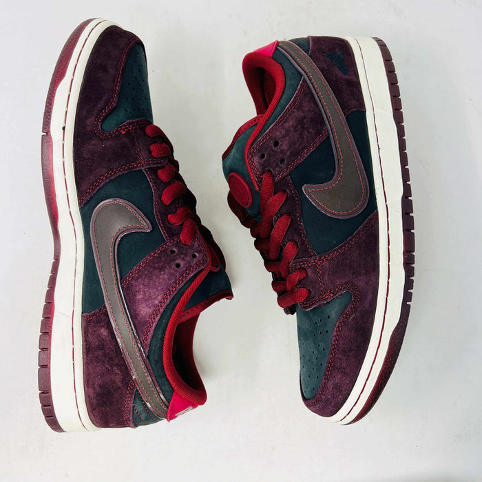 A BRAND NEW Nike SB Dunk Low Riot Skateshop sneaker featuring a black design with maroon accents, including a swoosh, laces, and a black sole with a white midsole, is displayed on top of its original white Nike SB shoebox.