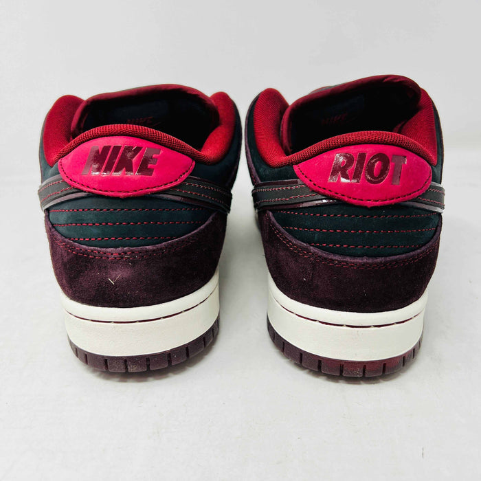 A BRAND NEW Nike SB Dunk Low Riot Skateshop sneaker featuring a black design with maroon accents, including a swoosh, laces, and a black sole with a white midsole, is displayed on top of its original white Nike SB shoebox.