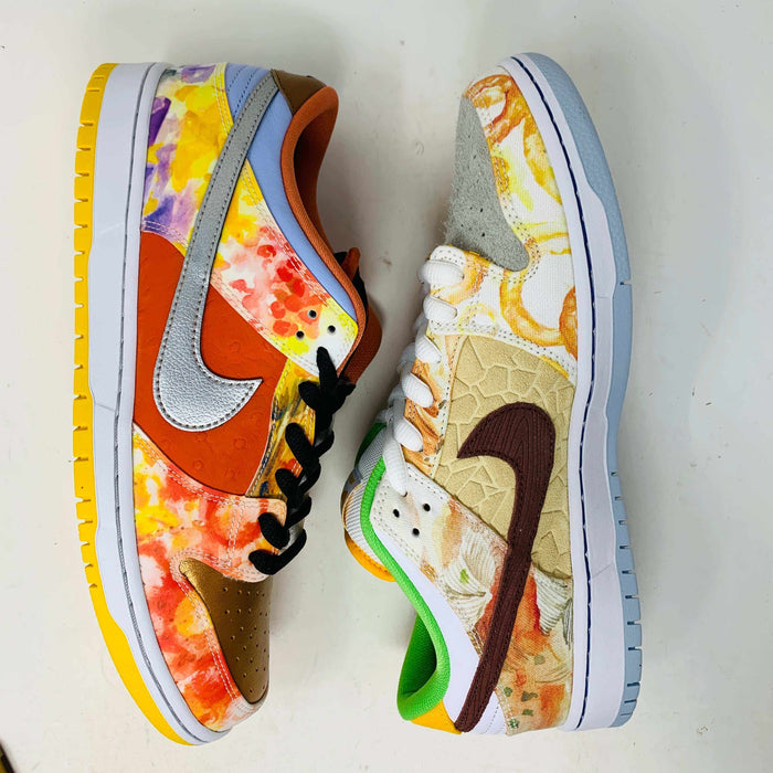 The Nike SB Dunk Low Street Hawker is a colorful sneaker displayed on a green and yellow box, featuring an authentic artistic design with brown, green, and orange tones. It has a quilted texture, white swoosh, white sole with light gray bottom, and comes with extra laces.