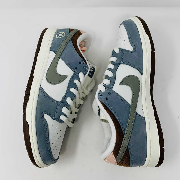 The Nike SB Dunk Low Yuto Horigome sneaker, with a white and light blue design featuring a gray swoosh, is displayed on a white Nike SB box. The authentic shoe has pink and brown laces, a brown sole, and an embroidered logo on the heel.