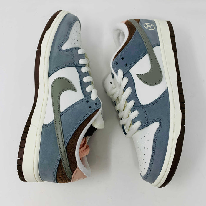 The Nike SB Dunk Low Yuto Horigome sneaker, with a white and light blue design featuring a gray swoosh, is displayed on a white Nike SB box. The authentic shoe has pink and brown laces, a brown sole, and an embroidered logo on the heel.