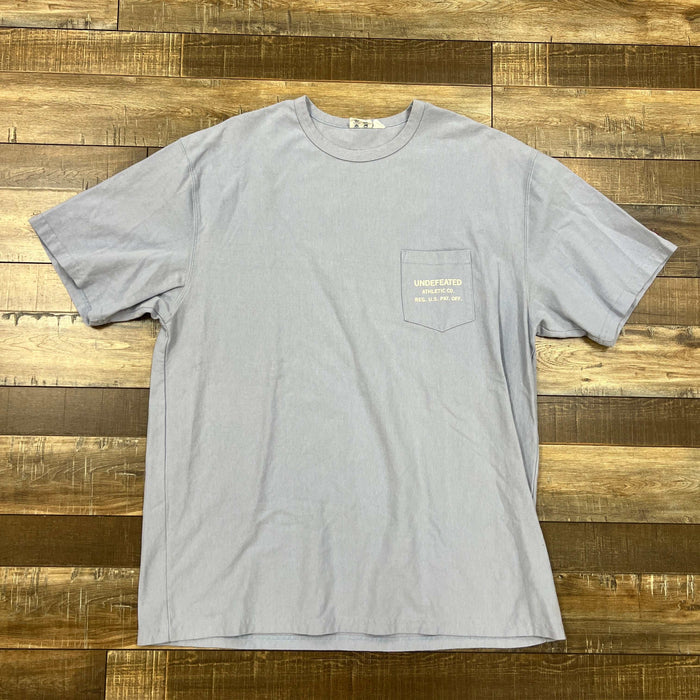 Undefeated Blue pocket tee shirt laid flat on wooden surface, preowned condition