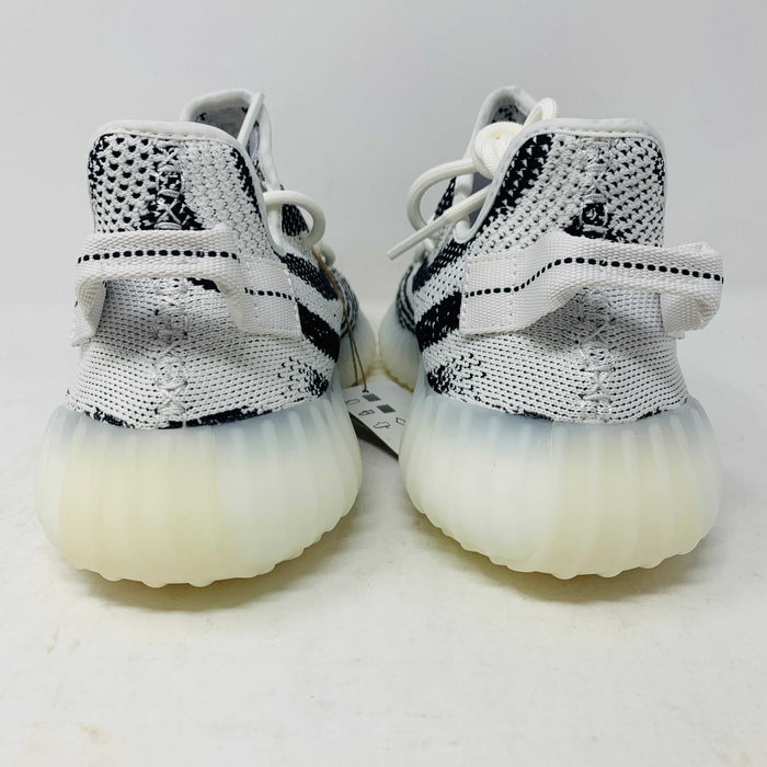 The Yeezy 350 V2 Zebra sneaker, 100% authentic, showcases SPLY-350 in bold red on a white and black design. Packaged in a box marked YZY 350 V2 MADE BY ADIDAS, it features a ribbed sole and knitted upper, effortlessly blending style and comfort.